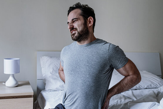 Mann sitzt auf einem Bett und hält sich schmerzhaft den Rücken
