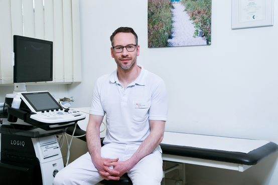 Arzt in weißer Berufskleidung sitzt in moderner Arztpraxis neben Ultraschallgerät und Untersuchungsliege